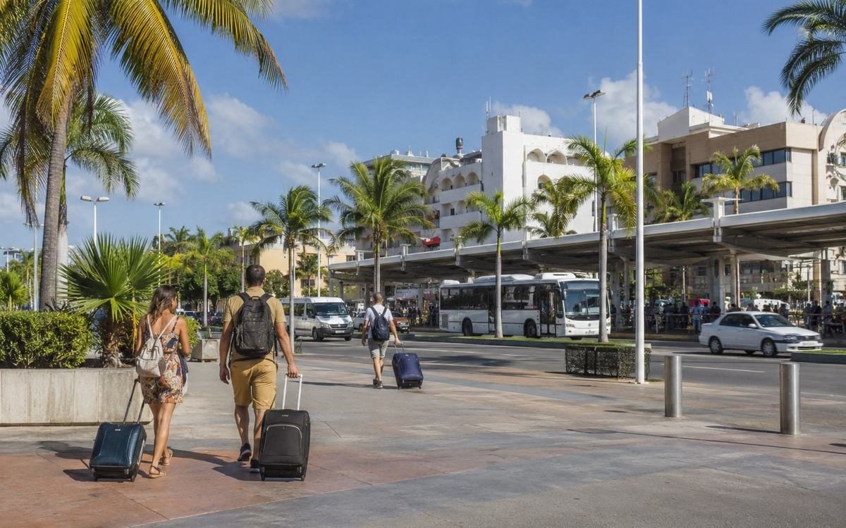 Travelers with rolling luggage arriving near a sunny downtown transit plaza.
