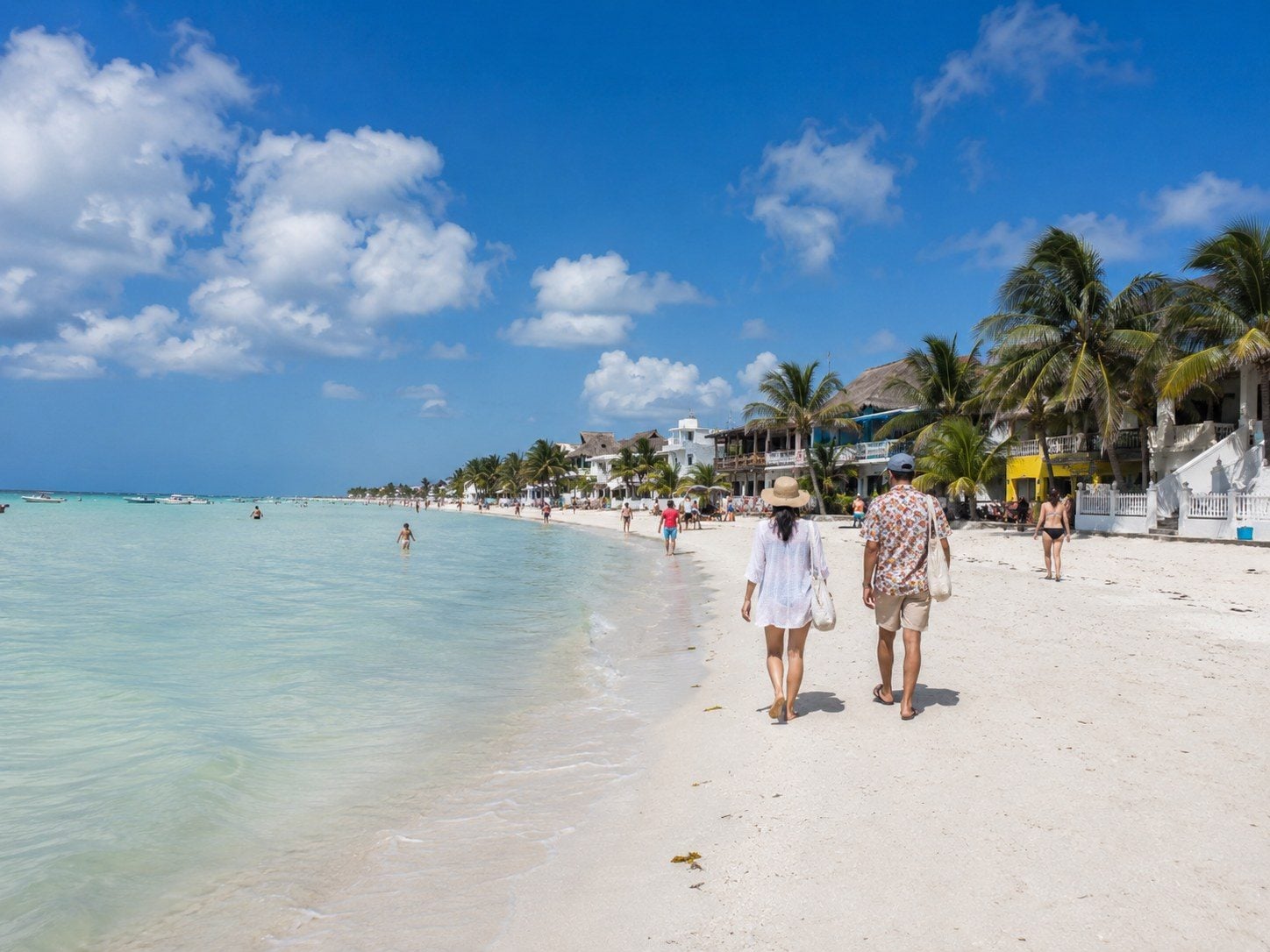 Visitors walking along Holbox Beach with shallow water and low-rise hotels in the distance.