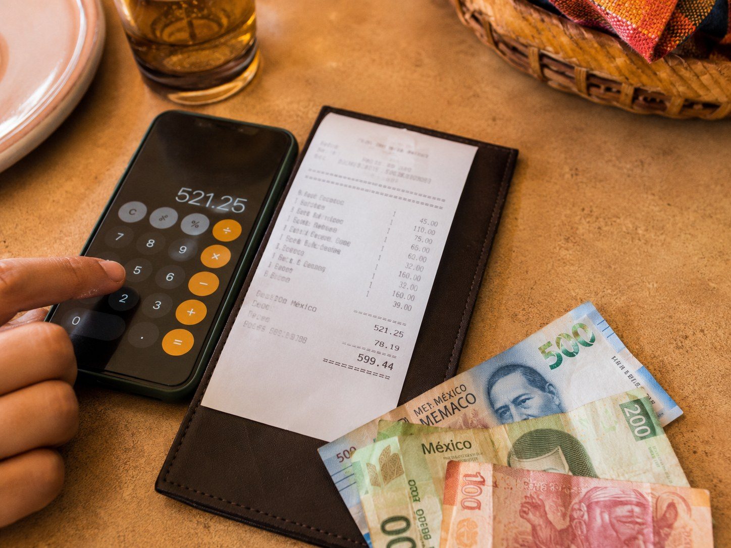Traveler using a phone calculator beside a restaurant bill and Mexican peso notes.